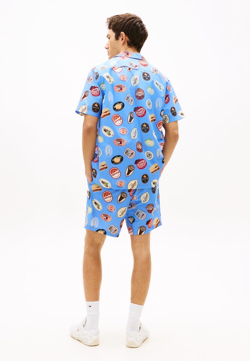 Homme debout, les mains dans les poches, portant une chemise à manches courtes et un short assortis de couleur bleu clair, couverts de logos circulaires colorés.