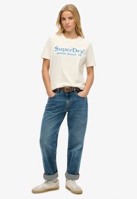 Kremowa bawełniana koszulka z niebieskim logo "SuperDry", w połączeniu z jeansami o wysokim stanie w kolorze niebieskim i białymi trampkami z beżowymi akcentami.