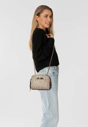 Beige Umhängetasche mit geprägtem Schriftzugmuster, schwarzem Logo, metallischen Beschlägen und einem schlanken verstellbaren Riemen. Getragen zu lässigem Denim und Pullover.