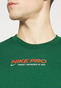 Hombre vistiendo una camiseta Nike Pro verde con texto del logo rojo y blanco, mostrando el cuello y la parte baja de la cara girados ligeramente hacia un lado.