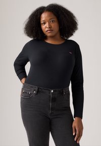 Mujer con cabello rizado natural, vistiendo una camiseta de manga larga negra y jeans oscuros de cintura alta, de pie contra un fondo claro y sencillo.