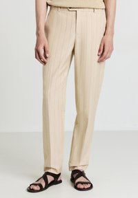 Beige gestreifte Hose aus leichtem Stoff, mit taillierter Passform, geraden Beinen und Seitentaschen, kombiniert mit dunkelbraunen Sandalen.