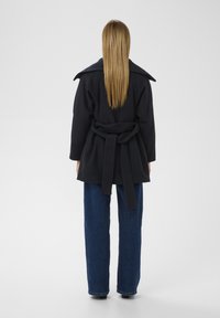 Tmavomodrý kabát s opaskom a širokým golierom, vyrobený z vlneného zmesi, s voľným strihom a rozparkom na chrbte. Kombinovaný s voľnými džínsovými nohavicami.