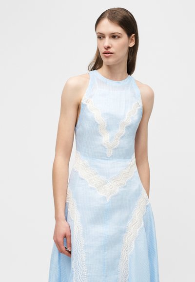 Νεαρή γυναίκα με αμάνικο ανοιχτό μπλε φόρεμα με λευκά, δαντελένια σχέδια, που κοιτάζει προς το πλάι, με απλό φόντο.