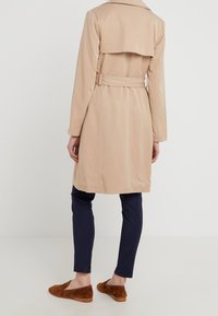 Beigefärgad trenchcoat med midjebälte, axelflikdetalj och en slät textur. Matchad med marinfärgade slim-fit byxor och bruna mockaloafers.