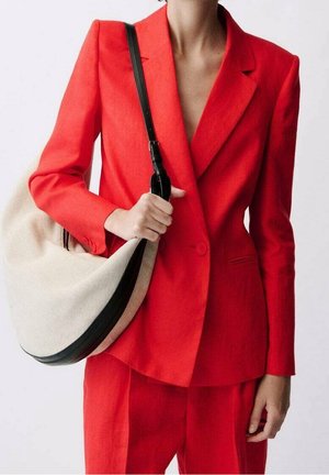 Persona con un traje rojo brillante sosteniendo un gran bolso beige y negro con una correa negra sobre un fondo liso.