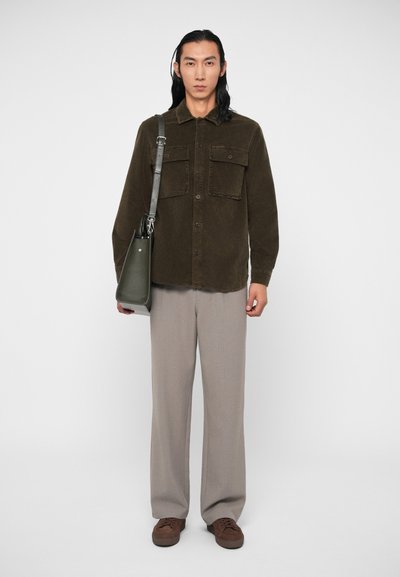 Camisa de veludo cotelê castanha com dois bolsos no peito, calças largas cinzas e sapatos castanhos. O conjunto inclui uma mala de ombro verde.