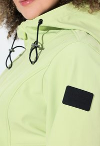 Veste vert clair avec fermeture éclair sur le devant, capuche à cordon de serrage et patch de marque noir sur la manche. Texture lisse et design ajusté.