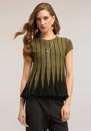 Kvinde med bølget hår iført olivengrøn top med sorte lodrette plisseringer, sorte bukser, perleøreringe med hæng, og sølv lariat-halskæde.