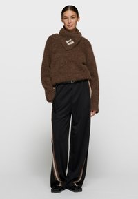 Suéter marrón peludo con cuello alto combinado con pantalones negros elásticos con franjas laterales. Modelo de pie sobre un fondo neutro.