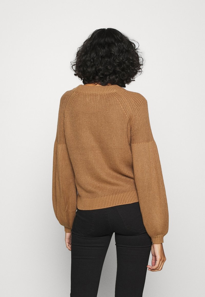 Femme aux cheveux bouclés portant un pull en maille marron avec des manches bouffantes et un pantalon noir, vue de dos sur un fond uni.