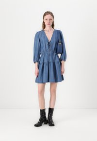 Džinsa kleita ar dziļu V-veida kakla izgriezumu, pūkainām piedurknēm, piegulošu jostu un izplestu svārku. Valkāta ar melniem potīšu zābakiem un zilu rokassomu.