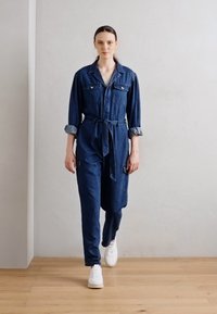 Une combinaison en denim bleu foncé, avec une taille nouée, des manches longues, deux poches poitrine et des poignets retroussés. Assortie à des baskets blanches.