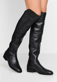 Bottes noires montantes en cuir lisse avec une coupe ajustée, semelle plate et panneaux latéraux élastiques pour le confort et la flexibilité.