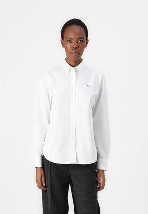 White long-sleeve button-up shirt with a classic collar and a small green crocodile logo on the chest. Fabric appears smooth and polished.