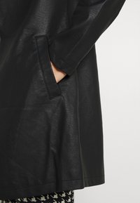 Trench de cuero negro largo con un acabado texturizado, que cuenta con un bolsillo lateral y un corte relajado. Leggings estampados visibles por debajo.