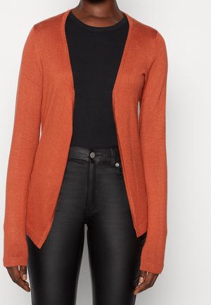 Persoon draagt een open roestoranje vest over een zwart shirt en zwarte broek met hoge taille tegen een effen lichte achtergrond.