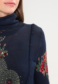 Marineblauer gestrickter Pullover mit einem Schalkragen, der mit Blumenstickerei und Pailletten verziert ist, sowie dekorativen lila Knöpfen entlang der Schulternaht.