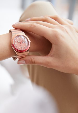 Montre à quartz Timex avec un cadran rose, des index horaires blancs et une lunette rouge. Elle possède un bracelet en métal avec des tons alternés de rose et d'or.