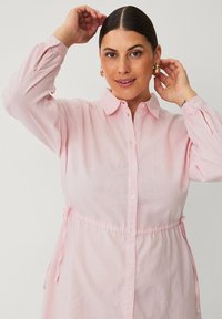 Lichtroze shirtjurk van zachte stof, met een knoopsluiting aan de voorkant, kraag en een cinched taille met strikjes. Lange mouwen met verstelbare manchetten.