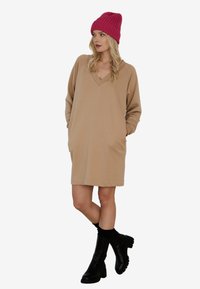 Beżowa sukienka z długim rękawem, dekoltem w kształcie litery V i bocznymi kieszeniami, w zestawieniu z różowym dzianinowym beretem i czarnymi botkami za kostkę. Gładka tekstura.