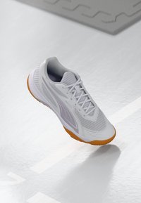 Białe i szare buty sportowe z pomarańczową podeszwą unoszące się nad gładką białą podłogą z oznaczeniami boiska sportowego.
