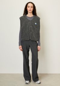 Melnā, teksturētā flīsa veste ar noapaļotu kakla izgriezumu, pogu aizdari un sarkanu logo. Valkāta virs garām piedurknēm pelēka tops un pelēkiem platiem biksēm.