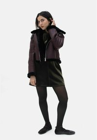 Burgunderfarbene Lederjacke in Cropped-Form mit schwarzem Kunstfellkragen, kombiniert mit einem dunkelgrünen Kleid. Das Modell trägt schwarze Strumpfhosen und Ballerinas.