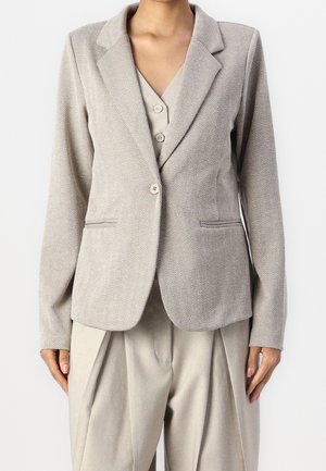 Blazer beige de espiga a medida con solapas con muesca y cierre de dos botones, usado sobre chaleco beige a juego y pantalones plisados y sueltos.