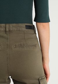 Pantalon cargo vert olive avec une taille cintrée et des poches arrière. Le tissu est lisse avec une texture mate, présentant une couture piquée et une étiquette sur la ceinture.