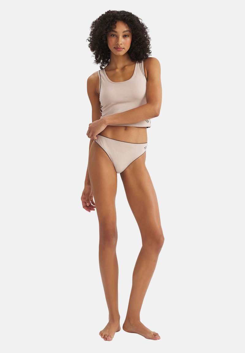 Femme debout pieds nus portant un crop top sans manches beige et une culotte taille haute assortie sur un fond blanc uni.