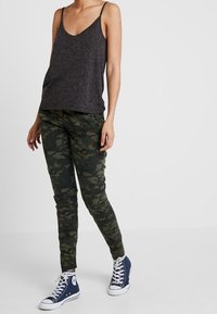 Černý pletený top bez rukávů s tenkými ramínky, zkombinovaný s upnutými zelenými maskáčovými kalhotami a modrými vysokými teniskami s bílými tkaničkami a podešvami.