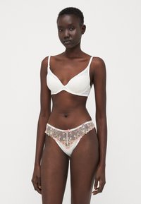 Weißes Spitzen-Bralette mit strukturiertem Finish und weichen Cups, kombiniert mit durchsichtigen weißen Panties mit floralen Stickereien und einem rosa Schleifenakzent.