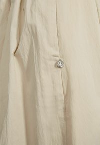 Beige plagg med en teksturert finish, med en sidelomme og en sirkulær sølvdetalj. Materialet er lett og lett krøllet.