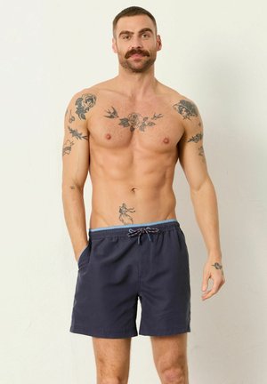 Hemdloser Mann mit Schnurrbart, der marineblaue Badeshorts trägt und Tätowierungen auf Brust, Armen und Bauch zeigt, vor schlichtem weißem Hintergrund stehend.