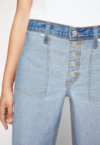 Zbliżenie jasnobłękitnych dżinsów z pięcioma widocznymi metalowymi guzikami oraz kontrastowym pomarańczowym przeszyciem, noszonych z białą bluzką i częściowo widoczną ręką.