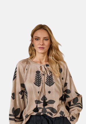 Donna bionda che indossa una blusa beige con grandi stampe floreali nere e lacci con nappine, abbinata a pantaloni neri, su sfondo semplice.