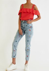 Czerwony top z odkrytymi ramionami ozdobiony falbanami, zestawiony z jasnoniebieskimi dżinsami z kolorową kwiatową haftem i białymi tenisówkami.