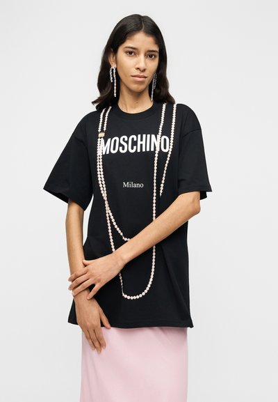 Μαύρο βαμβακερό μπλουζάκι με λευκό λογότυπο "MOSCHINO" και κείμενο "Milano", διακοσμημένο με λεπτομέρεια αλυσίδας με χάντρες· το μοντέλο φοράει ροζ φούστα.
