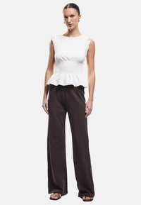 Witte peplumtop met smocktail en strikdetails op de schouders, gecombineerd met bruine wijde broek in een textuurstof. Zwarte sandalen.