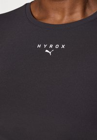 T-shirt de sport noir en tissu lisse, avec un col rond et le texte imprimé en blanc "HYROX" accompagné d'un petit logo Puma en dessous.