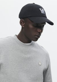 Black baseball cap with white "W" logo, worn with a gray sweatshirt featuring a round logo patch on the left chest. Smooth fabric texture.