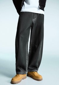 Černé široké džíny s hladkou texturou, s viditelným šitím, doplněné o béžovou obuv a bílou tričko pod černou bundou.