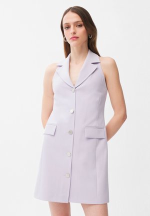 Donna che indossa un abito blazer lavanda senza maniche con bottoni argentati e tasche con patta, in piedi davanti a uno sfondo bianco.