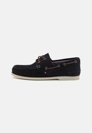 Suede navy boat shoe with a rounded toe, brown laces, and white rubber sole. Features contrasting stitching and a logo detail on the heel.