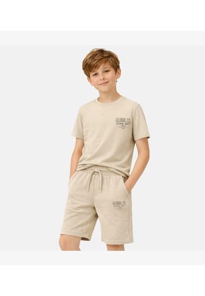 Jongen die een beige bijpassend T-shirt en short draagt met de tekst "Global Co. Denim Dept", staand met handen in de zakken, glimlachend tegen een witte achtergrond.