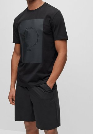Schwarzes Baumwoll-T-Shirt mit einem matten grafischen Design auf der Vorderseite. Kombiniert mit schwarzen Shorts, wodurch ein minimalistischer und monochromatischer Look entsteht.