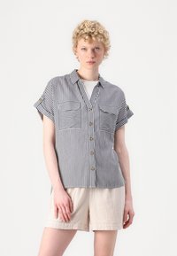 Striped short-sleeve shirt in black and white, featuring a button front, two chest pockets, and a relaxed fit. Paired with light beige shorts.