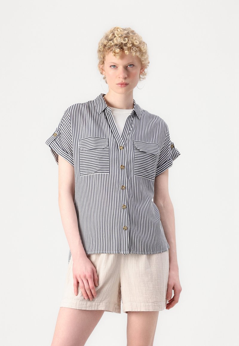 Striped short-sleeve shirt in black and white, featuring a button front, two chest pockets, and a relaxed fit. Paired with light beige shorts.