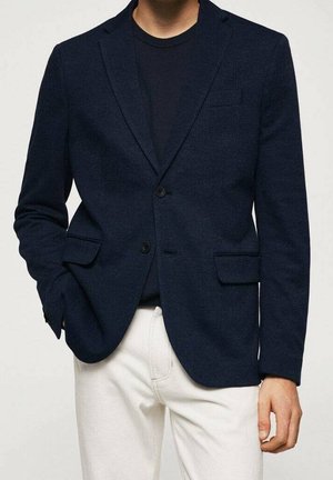 Homme portant un blazer à carreaux bleu marine foncé sur une chemise noire avec un pantalon blanc, une main dans la poche et l'autre détendue le long du corps.